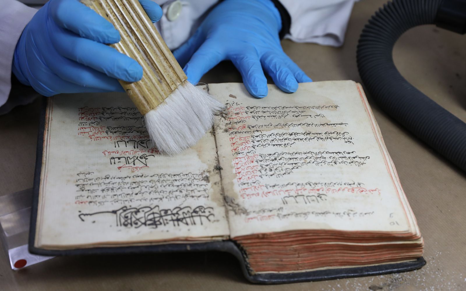 A conservator in blue gloves brushing dust from an open Maronite manuscript.